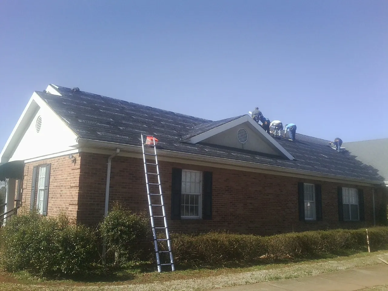 Crew tearing shingles off a brick ranch roof with a ladder set — Emergency Roof Repair work by our crew in Lucerne