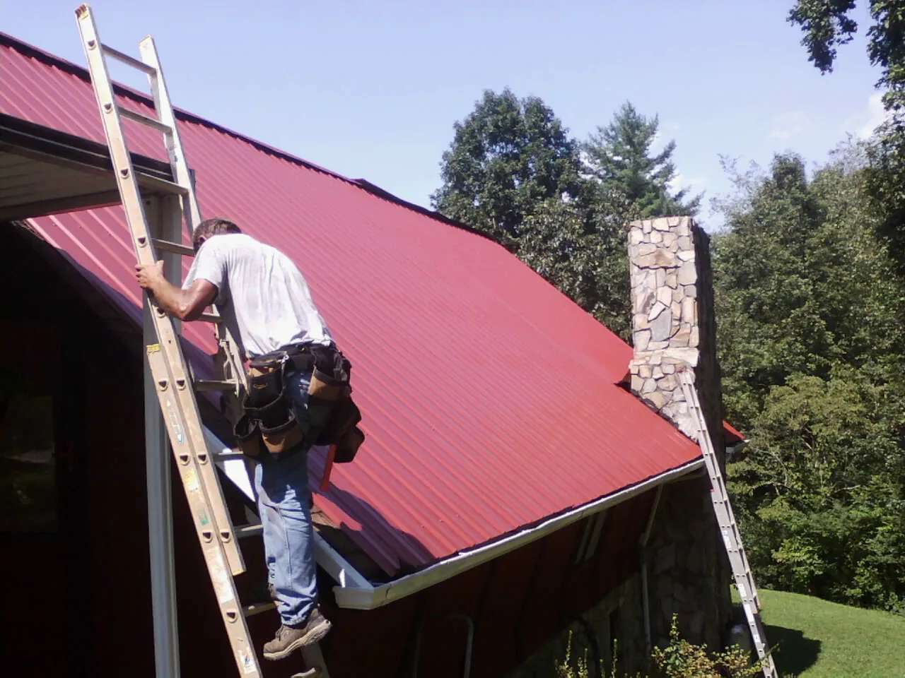 Roofer on a ladder beside a red metal roof and stone chimney — Soffit & Fascia Repair by Roofing Brothers in Lucerne